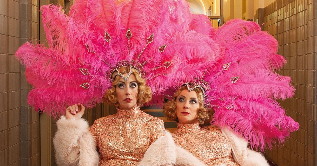 Promotiebeeld voor cabaretvoorstelling Harrekidee Plien en Bianca zitten naast elkaar en kijken bedenkelijk. Ze zijn gekleed in showgirl outfits: gouden glitterjurken, boa's en roze verentooien op hun hoofd. Promotiebeeld voor cabaretvoorstelling Harrekidee Plien en Bianca zitten naast elkaar en kijken bedenkelijk. Ze zijn gekleed in showgirl outfits: gouden glitterjurken, boa's en roze verentooien op hun hoofd.