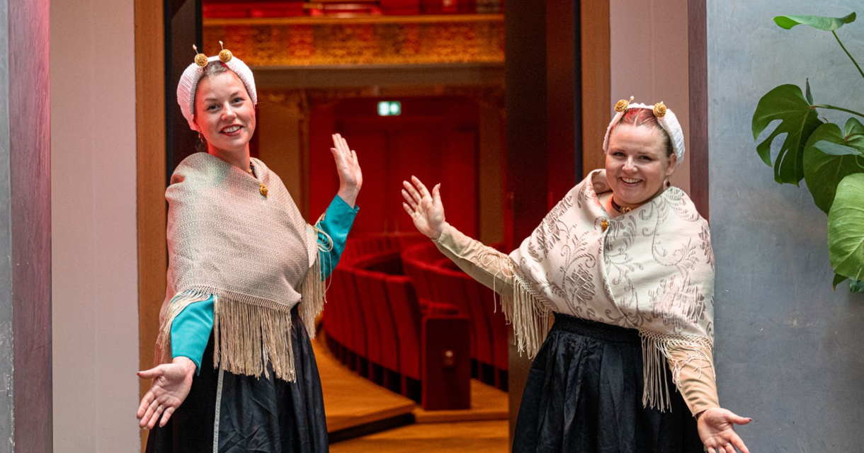 Twee vrouwen in traditionele klederdracht verwelkomen bezoekers bij een theateringang. Twee vrouwen in traditionele klederdracht verwelkomen bezoekers bij een theateringang.
