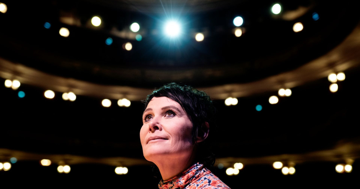 Vrouw op een toneel, belicht door een felle spot, met lege theaterstoelen op de achtergrond. Vrouw op een toneel, belicht door een felle spot, met lege theaterstoelen op de achtergrond.