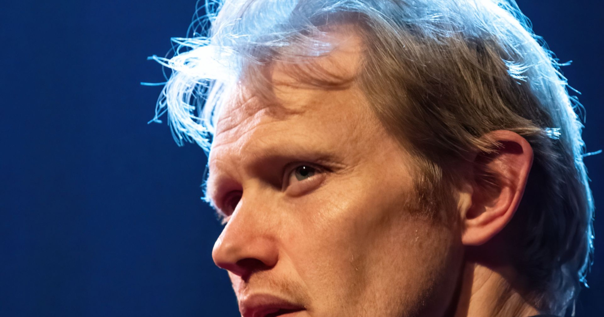 Close-up van een man met blond haar en serieuze blik, dramatisch belicht tegen een donkere achtergrond met blauw licht. Close-up van een man met blond haar en serieuze blik, dramatisch belicht tegen een donkere achtergrond met blauw licht.