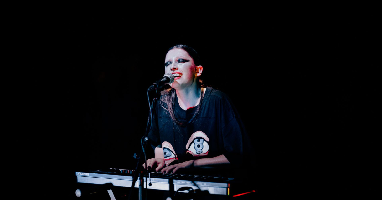 Artiest met opvallende make-up speelt keyboard en zingt op een donker podium, verlicht door een spotlight. Artiest met opvallende make-up speelt keyboard en zingt op een donker podium, verlicht door een spotlight.