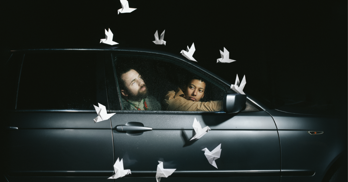 Twee personen zitten in een stilstaande auto terwijl witte papieren duiven rondom hen lijken te zweven in het donker Twee personen zitten in een stilstaande auto terwijl witte papieren duiven rondom hen lijken te zweven in het donker
