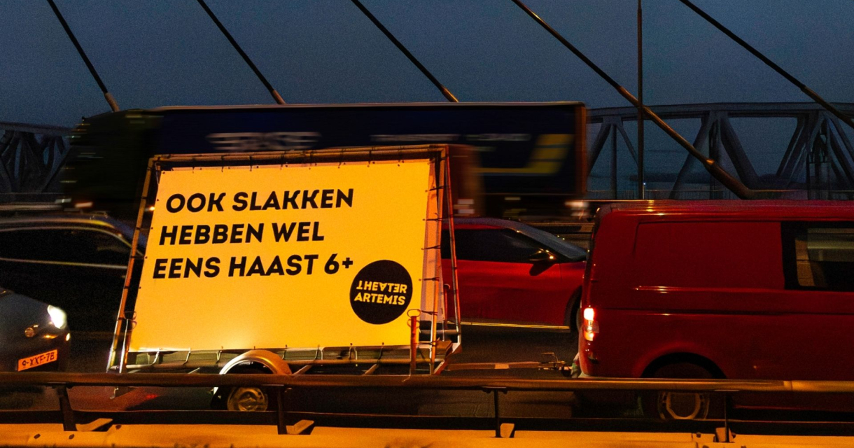 Verlichte aanhanger met tekst “Ook slakken hebben wel eens haast 6+” tussen stilstaand verkeer op brug in de avondspits