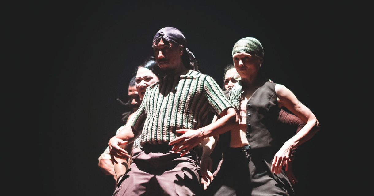 Groep dansers op een donker podium, intens belicht, met bandana’s en streetwear in een dynamische pose