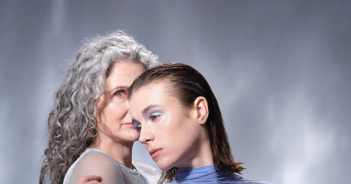 Twee vrouwen omarmen elkaar, één met grijs krullend haar en één met glad, donker haar, voor een grijze achtergrond. Twee vrouwen omarmen elkaar, één met grijs krullend haar en één met glad, donker haar, voor een grijze achtergrond.