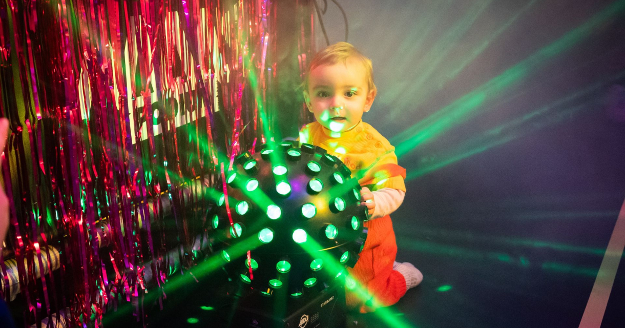 Peuter in oranje outfit kruipt bij discobal met groene lichtstralen en roze slierten op de achtergrond Peuter in oranje outfit kruipt bij discobal met groene lichtstralen en roze slierten op de achtergrond