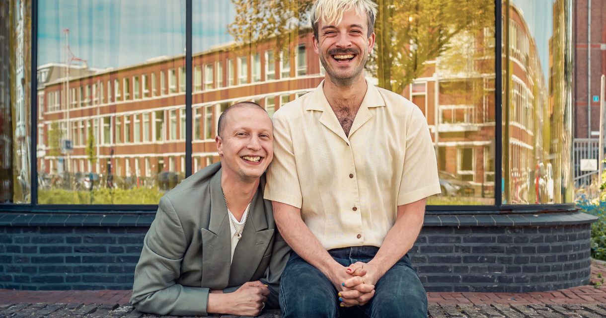 Twee lachende mannen zitten op een bakstenen muurtje voor een spiegelend raam met een stadspand op de achtergrond. De man links draagt een grijs pak, de man rechts een crèmekleurig overhemd en heeft blond haar. Twee lachende mannen zitten op een bakstenen muurtje voor een spiegelend raam met een stadspand op de achtergrond. De man links draagt een grijs pak, de man rechts een crèmekleurig overhemd en heeft blond haar.