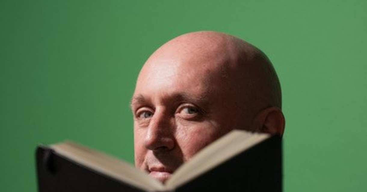 Kale man kijkt over een geopend boek richting camera, met groene achtergrond Kale man kijkt over een geopend boek richting camera, met groene achtergrond