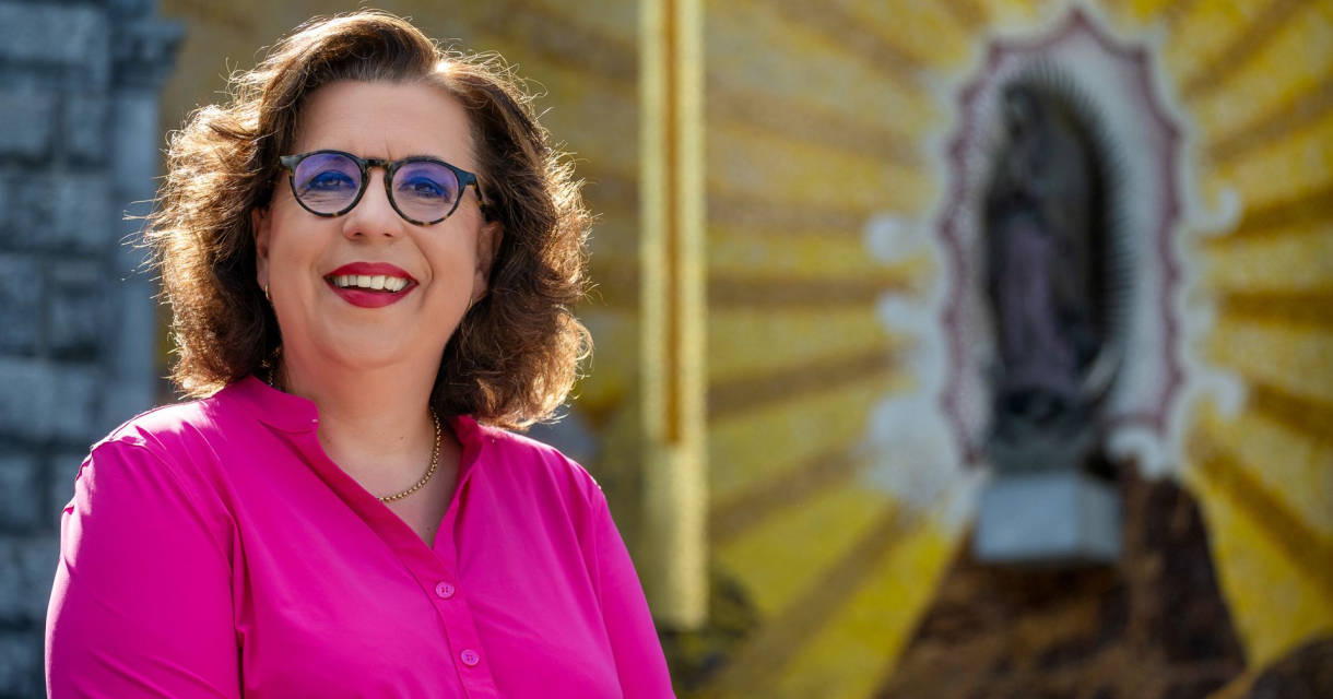 Vrouw met bril en felroze blouse lacht voor muurschildering met heiligenfiguur en stralend geel patroon