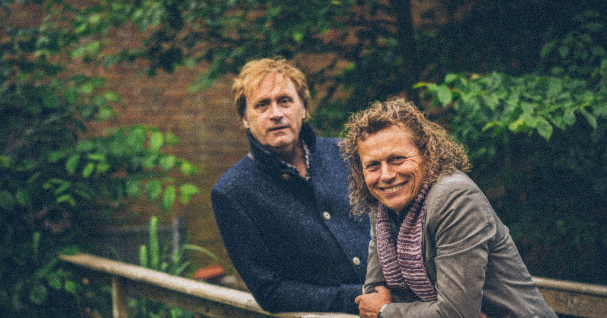 Syb van der Ploeg en Maarten Peters leunen over de reling van een brug omringt door bomen. Syb van der Ploeg en Maarten Peters leunen over de reling van een brug omringt door bomen.