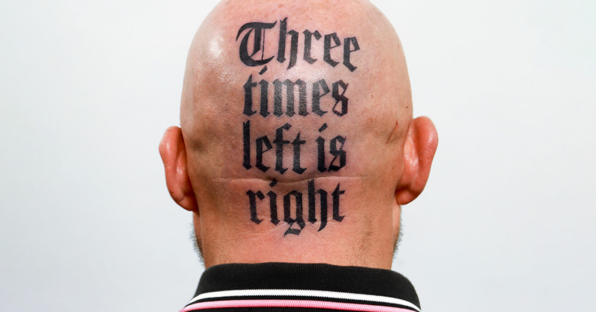 Kaalgeschoren achterhoofd van een persoon met een opvallende tatoeage in gotisch lettertype waarop staat: "Three times left is right". Kaalgeschoren achterhoofd van een persoon met een opvallende tatoeage in gotisch lettertype waarop staat: "Three times left is right".