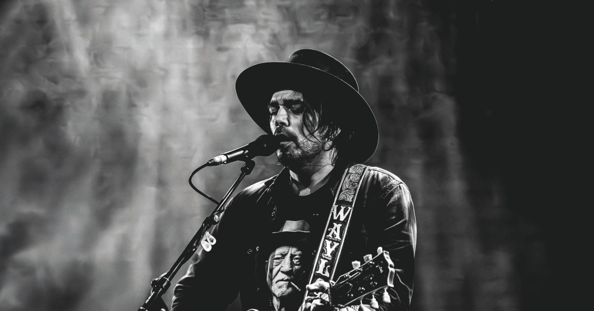 Zwart-witfoto van een man met hoed die zingt en gitaar speelt op een podium, met een expressieve blik en een shirt met een portret erop. Zwart-witfoto van een man met hoed die zingt en gitaar speelt op een podium, met een expressieve blik en een shirt met een portret erop.
