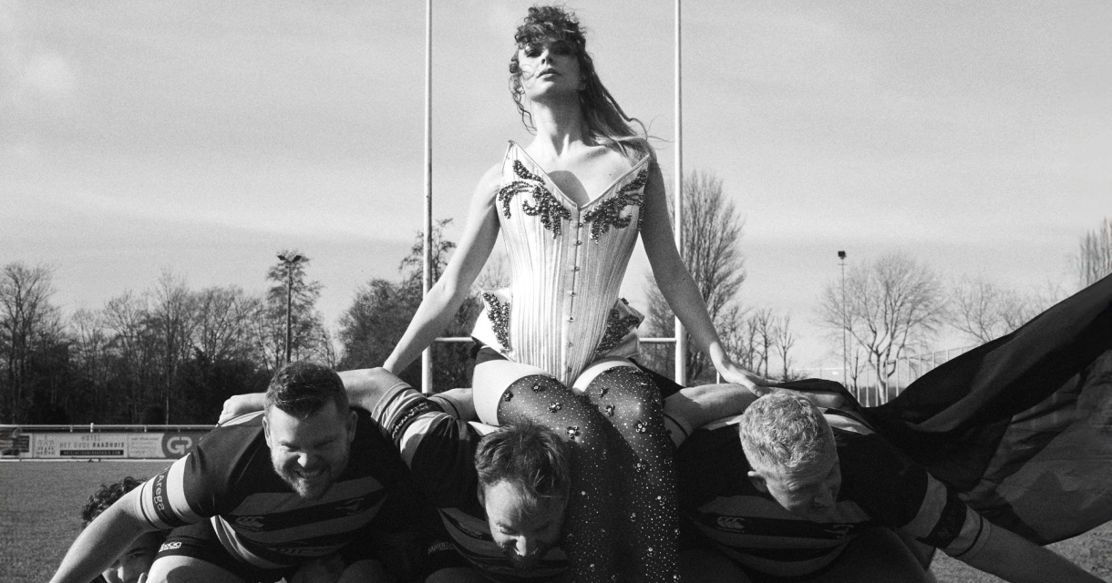 Zangeres in glitteroutfit zit op rugbyscrum van drie mannen met Superman-shirts op een sportveld Zangeres in glitteroutfit zit op rugbyscrum van drie mannen met Superman-shirts op een sportveld