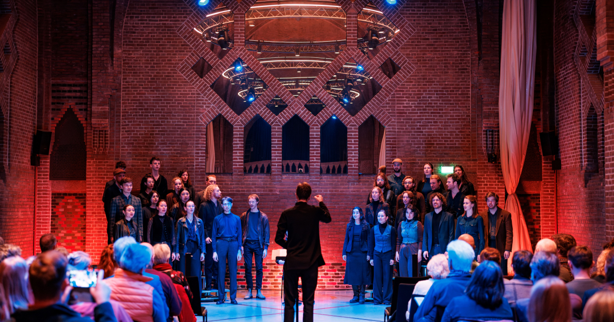 Het Nederlands Studenten Kamerkoor voert een concert uit in een sfeervol verlichte kerk, onder leiding van een dirigent. Het Nederlands Studenten Kamerkoor voert een concert uit in een sfeervol verlichte kerk, onder leiding van een dirigent.