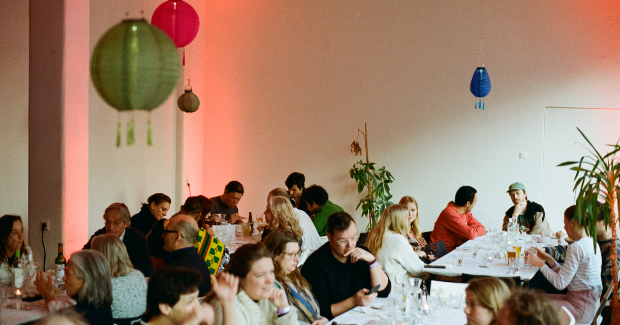 Gezellige groepsbijeenkomst in een sfeervol verlichte ruimte, waar mensen aan lange tafels eten, drinken en met elkaar in gesprek zijn. Kleurrijke lampions en planten zorgen voor een informele, warme sfeer. Gezellige groepsbijeenkomst in een sfeervol verlichte ruimte, waar mensen aan lange tafels eten, drinken en met elkaar in gesprek zijn. Kleurrijke lampions en planten zorgen voor een informele, warme sfeer.