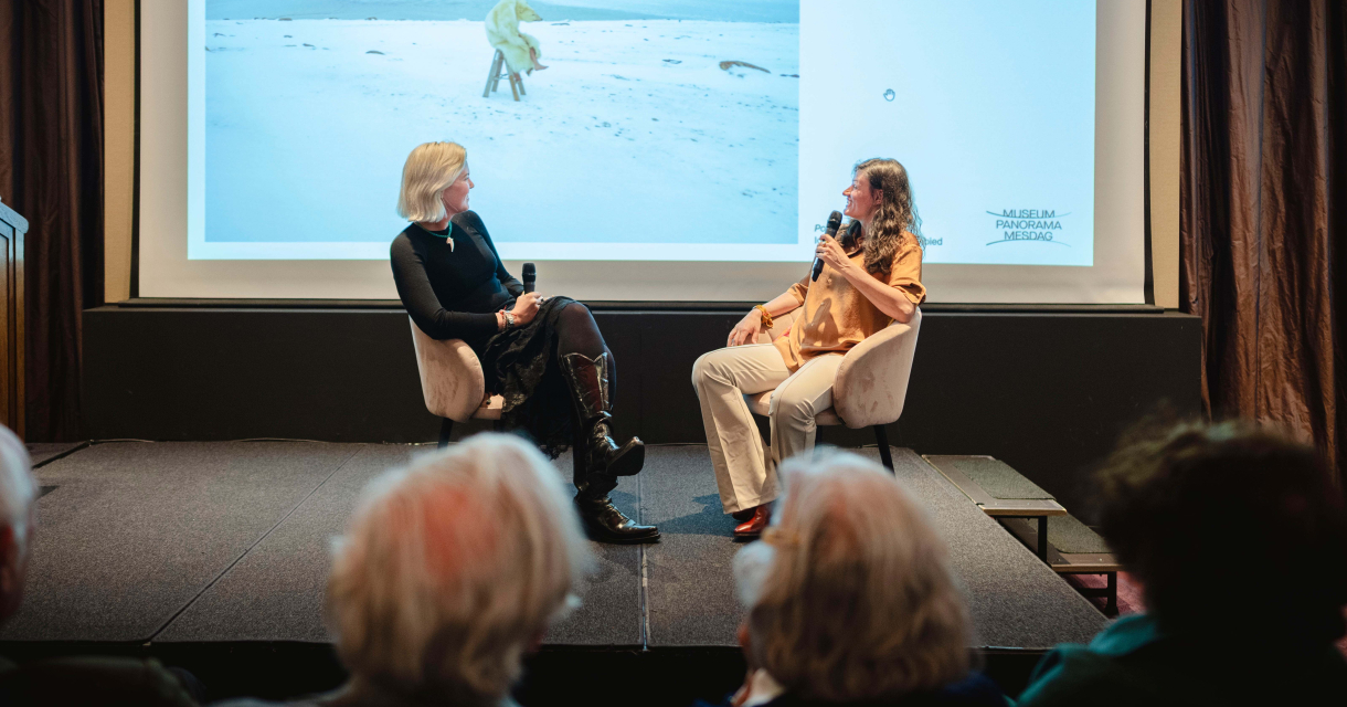 Twee vrouwen in gesprek op een podium met een projectie van een poollandschap op de achtergrond, terwijl het publiek toekijkt. Twee vrouwen in gesprek op een podium met een projectie van een poollandschap op de achtergrond, terwijl het publiek toekijkt.