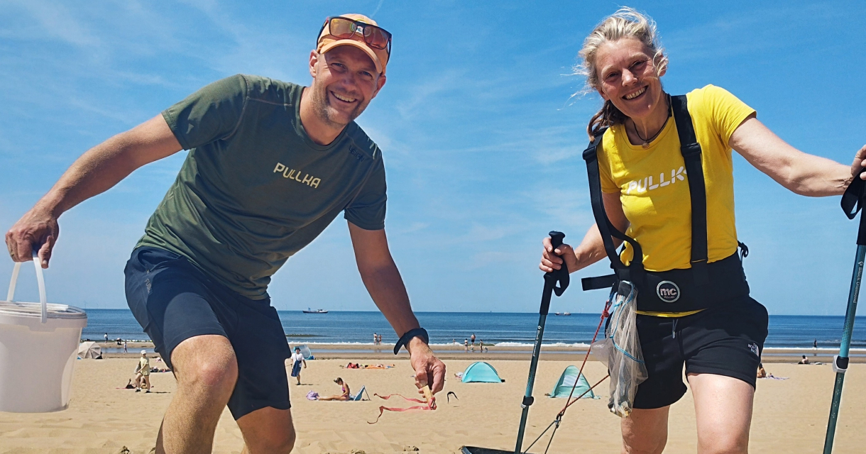 Twee mensen verzamelen afval op het strand met een emmer en grijper, op de achtergrond zee en strandtenten.
