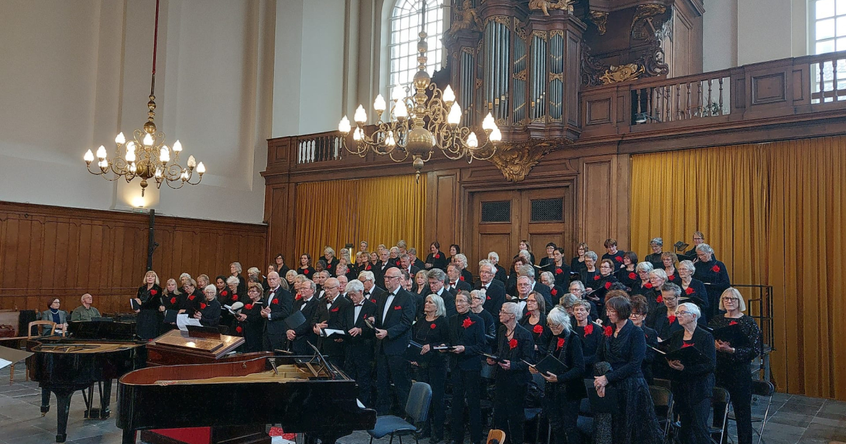 Groot koor in zwarte kleding zingt in een kerkzaal met houten lambrisering en kroonluchters