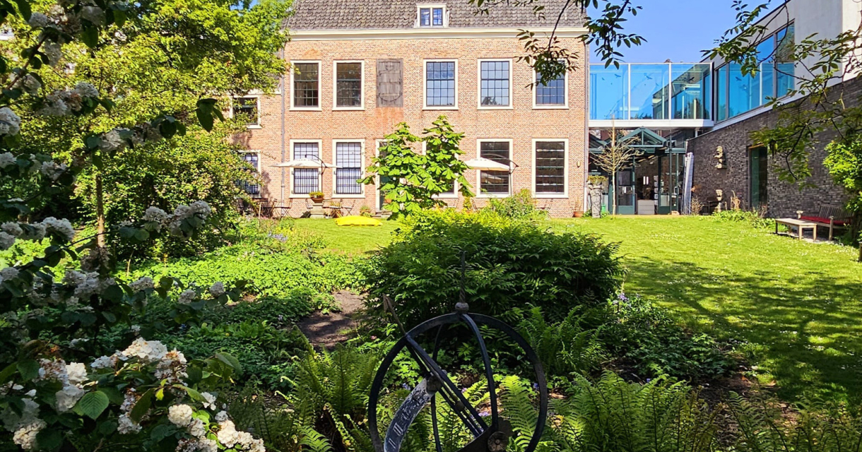 Een groene binnentuin met bloemen, struiken en een decoratief zonnewijzerbeeld, met op de achtergrond een historisch gebouw met luiken en een moderne glazen aanbouw aan de rechterzijde.