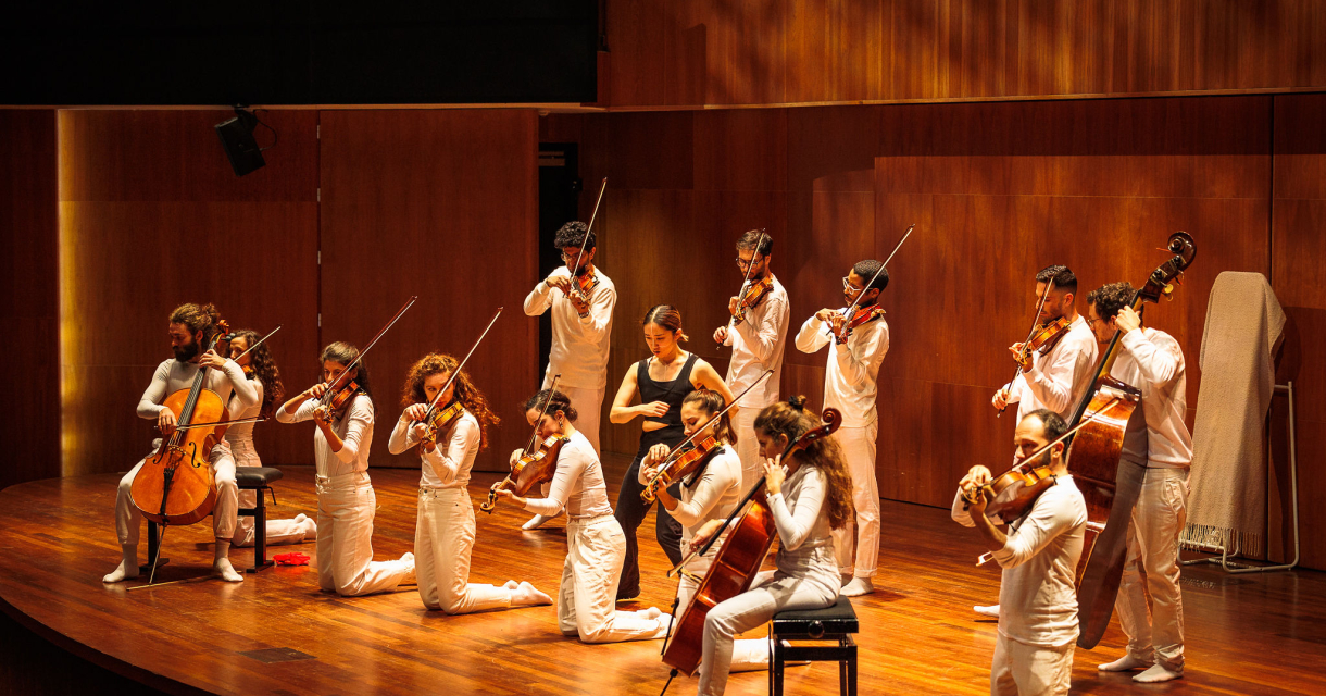 Strijkensemble in witte outfits voert een dynamisch optreden uit op een houten podium, met musici zittend, staand en knielend terwijl ze spelen op violen, altviolen en cello’s. Strijkensemble in witte outfits voert een dynamisch optreden uit op een houten podium, met musici zittend, staand en knielend terwijl ze spelen op violen, altviolen en cello’s.