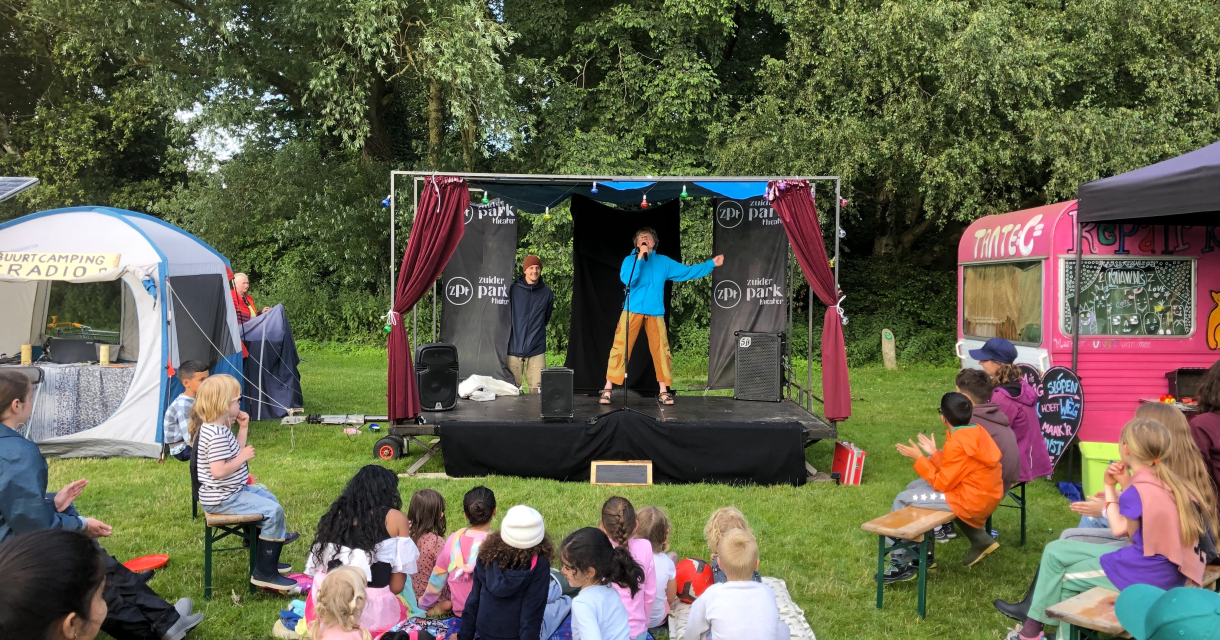 Kindervoorstelling in de buitenlucht met een klein podium tussen gekleurde caravans. Een performer spreekt het publiek toe, terwijl kinderen en ouders op kleedjes en bankjes toekijken op het gras. Kindervoorstelling in de buitenlucht met een klein podium tussen gekleurde caravans. Een performer spreekt het publiek toe, terwijl kinderen en ouders op kleedjes en bankjes toekijken op het gras.
