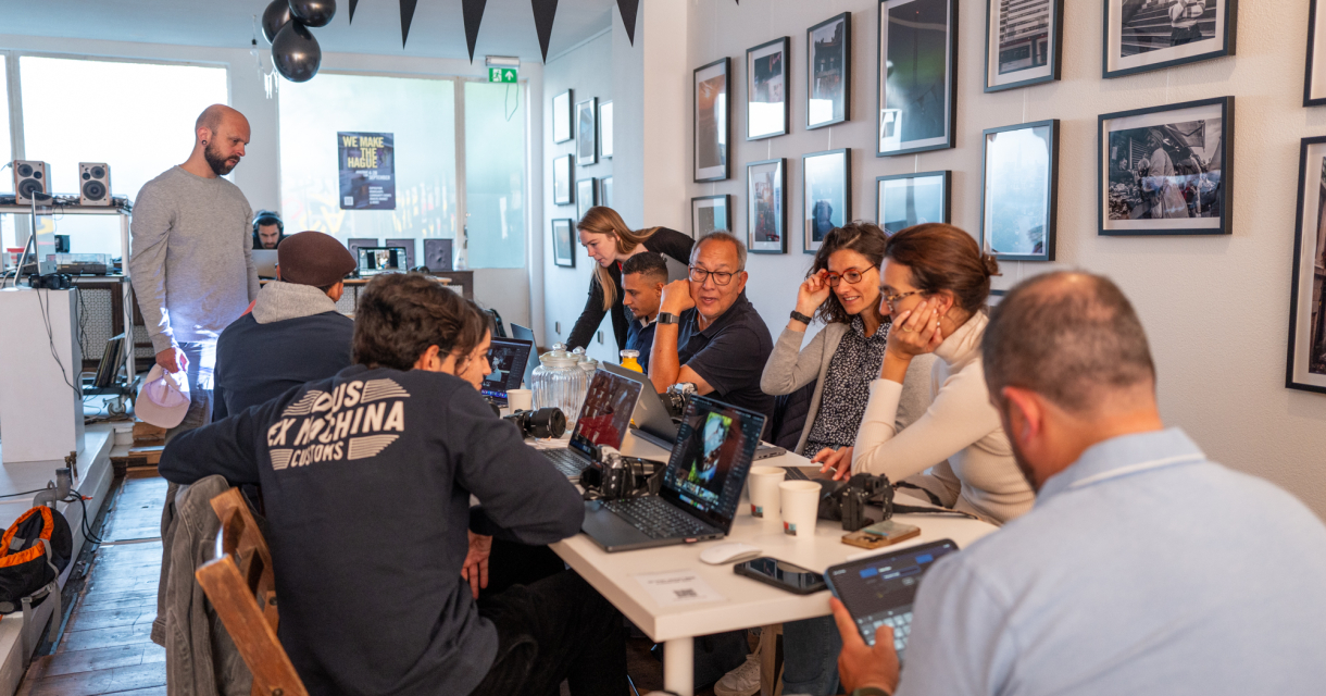 Groep mensen volgt een fotografieworkshop aan een lange tafel, terwijl een man uitleg geeft in een creatief ingerichte ruimte met foto’s aan de muur. Groep mensen volgt een fotografieworkshop aan een lange tafel, terwijl een man uitleg geeft in een creatief ingerichte ruimte met foto’s aan de muur.