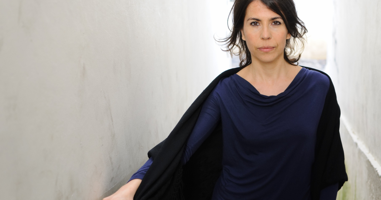 Vrouw met donker haar in een smalle gang, gekleed in een donkerblauwe top en zwarte omslagdoek, kijkt recht in de camera met een serieuze blik. Vrouw met donker haar in een smalle gang, gekleed in een donkerblauwe top en zwarte omslagdoek, kijkt recht in de camera met een serieuze blik.