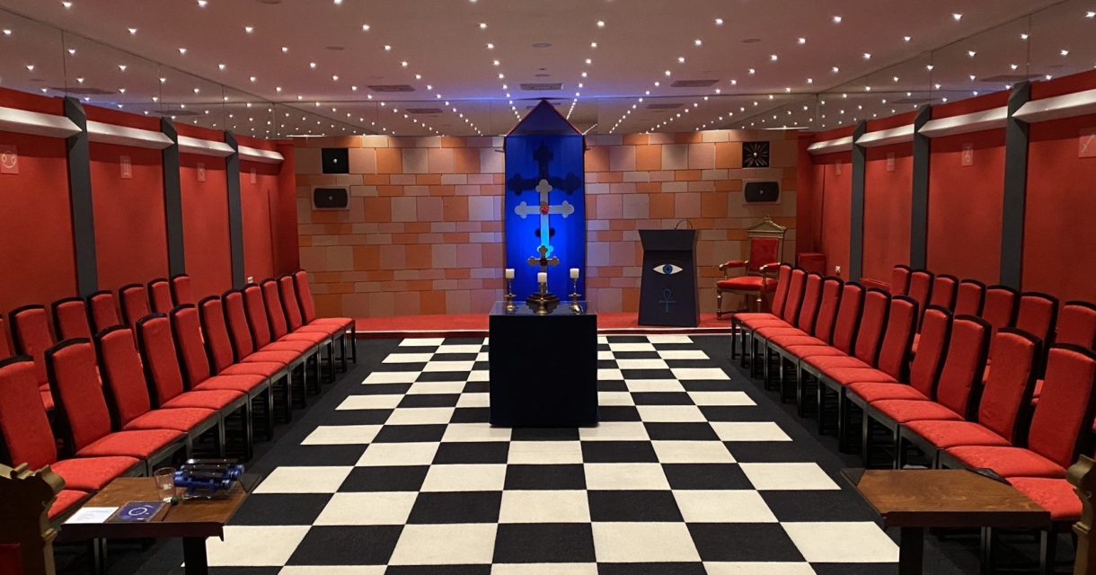 Symmetrisch ingerichte zaal met rood gestoffeerde stoelen langs de wanden, een zwart-wit geblokte vloer en een centraal altaar met blauwe verlichting. Symmetrisch ingerichte zaal met rood gestoffeerde stoelen langs de wanden, een zwart-wit geblokte vloer en een centraal altaar met blauwe verlichting.