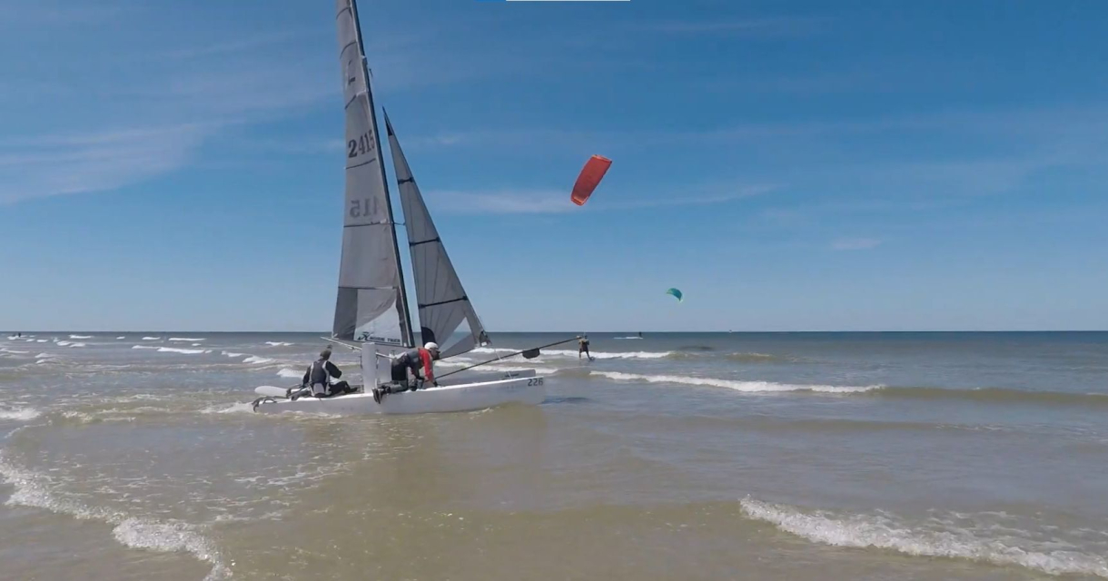 Catamaran met opgestoken zeilen vaart de zee op, met kitesurfers verderop in beeld onder blauwe lucht Catamaran met opgestoken zeilen vaart de zee op, met kitesurfers verderop in beeld onder blauwe lucht