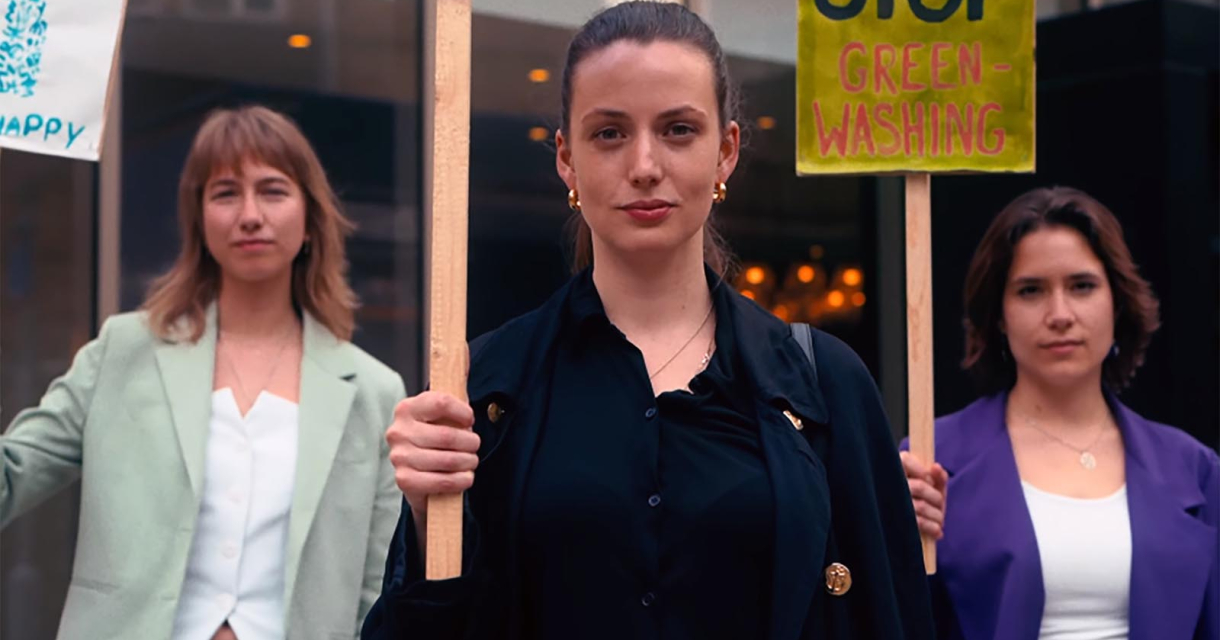 Drie jonge vrouwen houden protestborden vast tijdens een demonstratie. De vrouw in het midden kijkt vastberaden in de camera. Eén van de borden heeft de tekst "Stop greenwashing". Ze staan voor een modern gebouw. Drie jonge vrouwen houden protestborden vast tijdens een demonstratie. De vrouw in het midden kijkt vastberaden in de camera. Eén van de borden heeft de tekst "Stop greenwashing". Ze staan voor een modern gebouw.