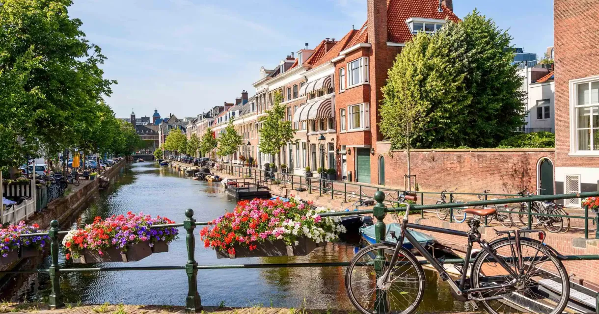 Zonnig straatbeeld in een Nederlandse stad met een gracht, bloemen aan de brugleuning en een geparkeerde fiets op de voorgrond. Zonnig straatbeeld in een Nederlandse stad met een gracht, bloemen aan de brugleuning en een geparkeerde fiets op de voorgrond.