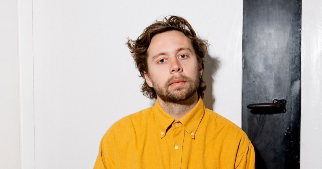 Een jonge man met halflang haar en een baard kijkt neutraal in de camera. Hij draagt een felgele overhemd en zit tegen een witte muur met een zwarte deur of paneel aan de rechterkant. Een jonge man met halflang haar en een baard kijkt neutraal in de camera. Hij draagt een felgele overhemd en zit tegen een witte muur met een zwarte deur of paneel aan de rechterkant.