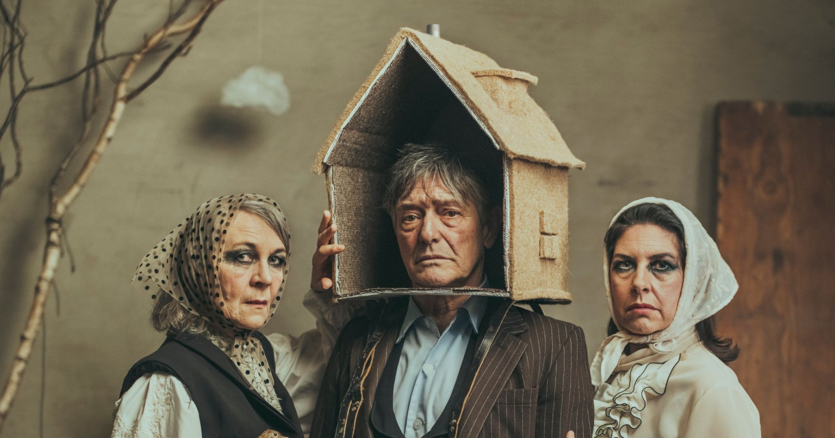 Drie mensen in theatrale kostuums poseren met serieuze gezichten; de persoon in het midden draagt een huis als hoofddeksel. Drie mensen in theatrale kostuums poseren met serieuze gezichten; de persoon in het midden draagt een huis als hoofddeksel.