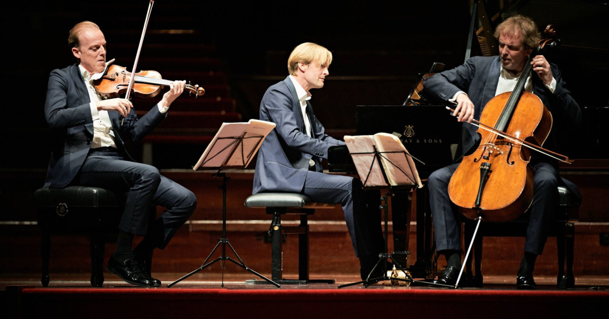Drie muzikanten voeren een klassiek pianotrio uit op een concertpodium, met viool, piano en cello. Drie muzikanten voeren een klassiek pianotrio uit op een concertpodium, met viool, piano en cello.