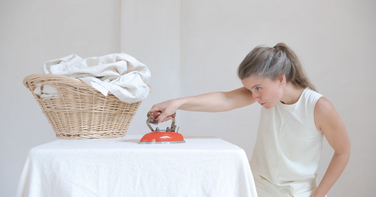 Vrouw in witte outfit strijkt geconcentreerd een tafelkleed met een klein oranje strijkijzer naast een rieten wasmand