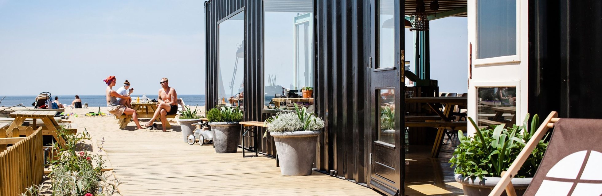 Mensen lekker genietend van de zon op het zij-terras van The Shore op Scheveningen. Deze duurzame strandtent bevindt zich in een container.