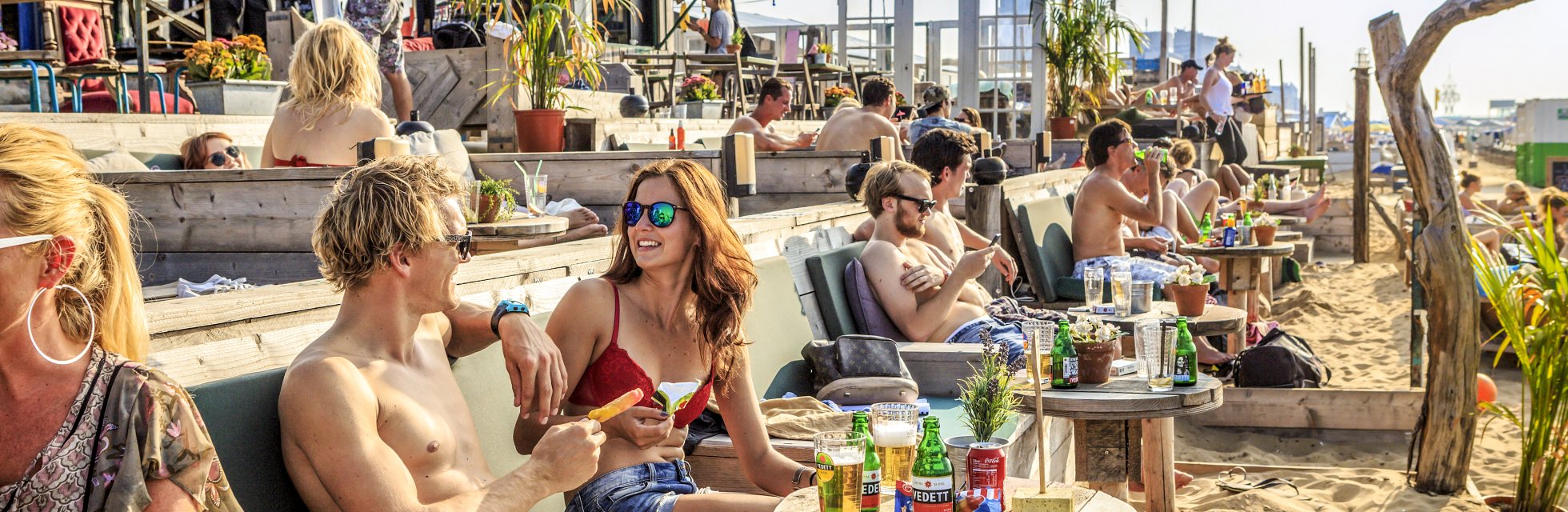 Een gezellig druk terras met mensen in badkleding met hun voeten in het zand bij strandtent Indigo op Scheveningen in Den Haag