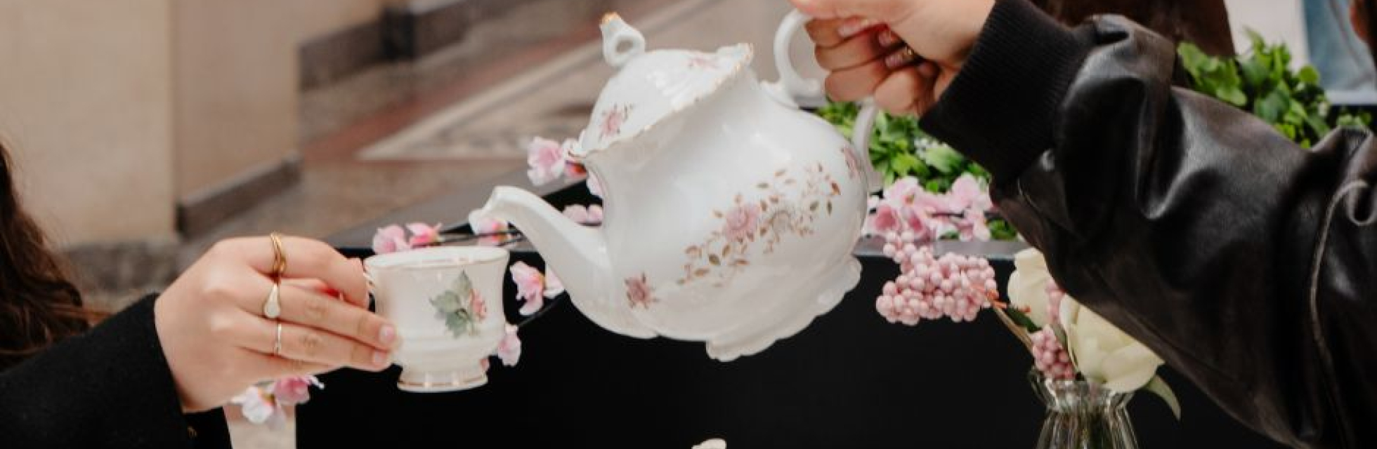 Twee mensen genieten van een high tea aan een sfeervol gedekte tafel in De Passage, met porseleinen servies, luxe taartjes en chocolade, terwijl thee wordt ingeschonken.