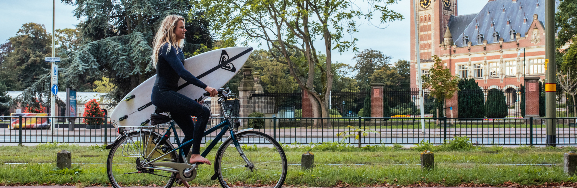 Zomer in Den Haag. Meisje met surfplank onder haar arm fiets langs het Vredespaleis richting Scheveningen