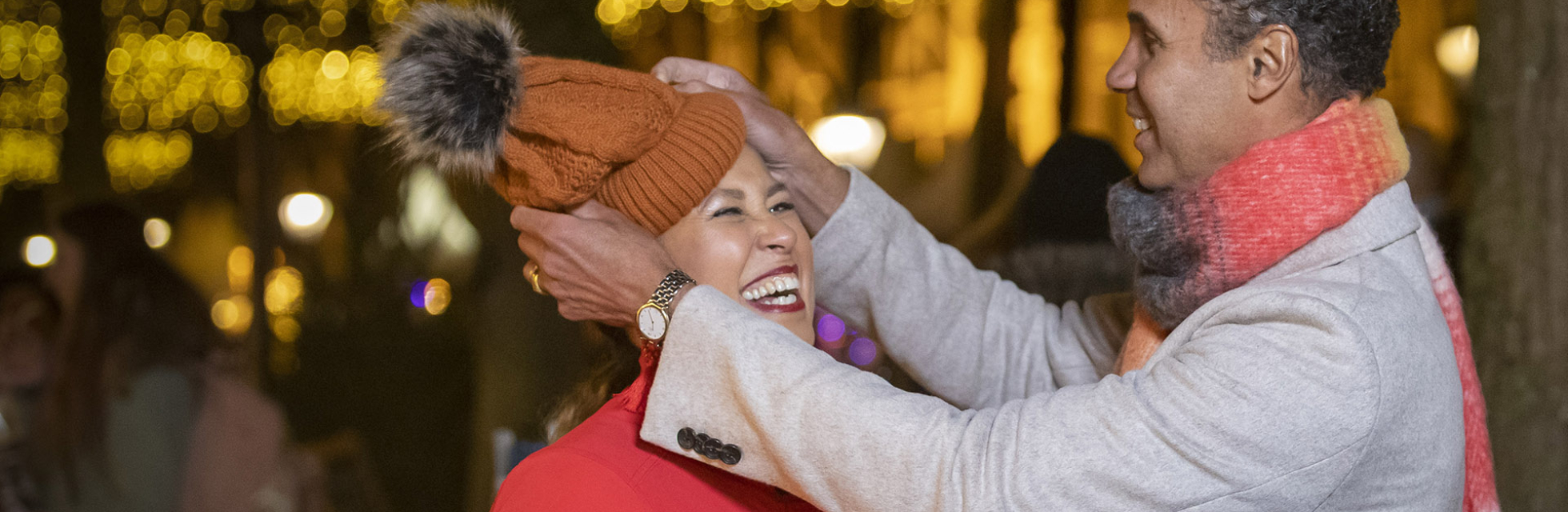 A woman laughs as a man places an orange hat on her head, surrounded by festive lights.