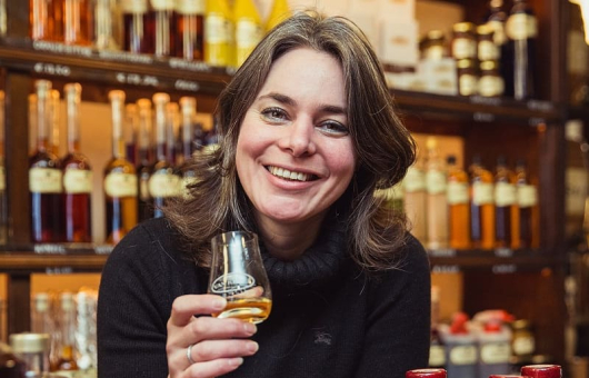 Fleur Kruyt met glas Van Kleef likeur aan de counter voor een schap flessen