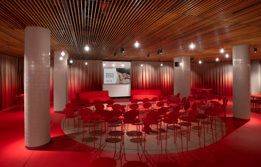 Zaalopstelling met rode stoelen in halve cirkel, podium met scherm, houten plafond en pilaren in modern interieur
