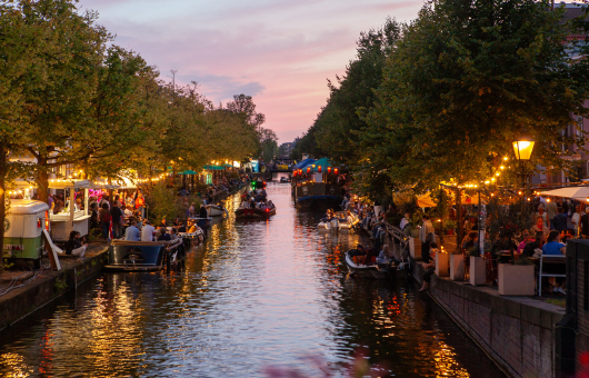 Gracht bij zonsondergang met volle terrassen, verlichte bomen en boten vol mensen tijdens een avondfestival