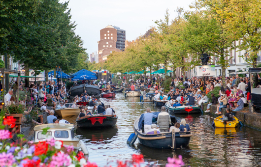 Drukke gracht vol boten met mensen, publiek zit langs de kade tussen bomen en terrassen in zomerse sfeer