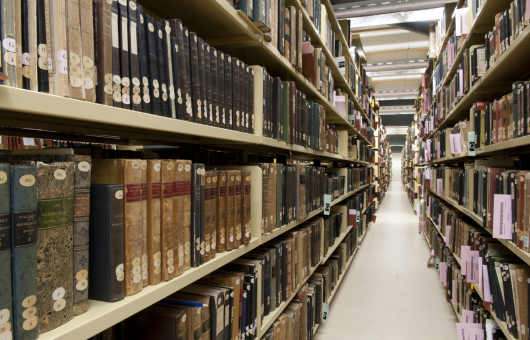 Smalle gang tussen hoge archiefrekken vol oude boeken en gebonden tijdschriften in depotomgeving