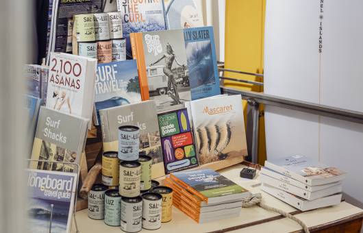 Een tafel met surfboeken, blikken en surfplanken op de achtergrond.