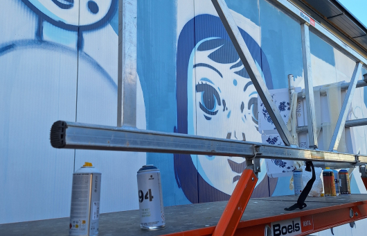 Een muurschildering van een gezicht in blauwe en witte kleuren, met verfspuitbussen op een ladder.