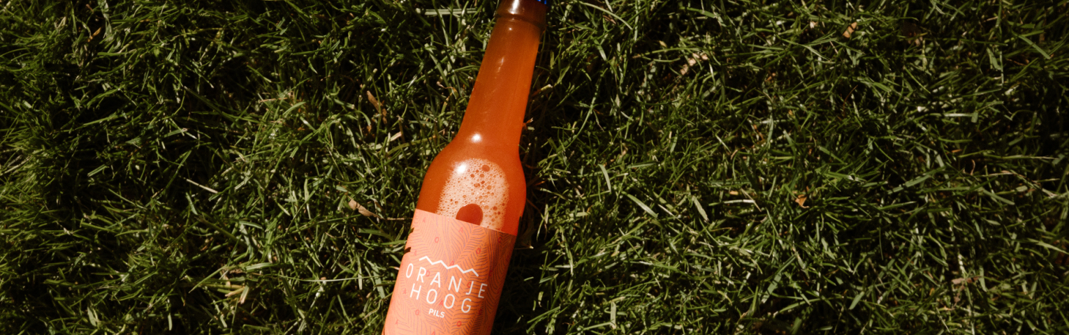 Een fles Oranje Hoog pils ligt op het gras.