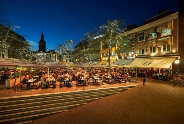De terrassen op de Grote markt gezellig verlicht in de avond. Op de achtergrond het silhouet van de Grote Kerk.