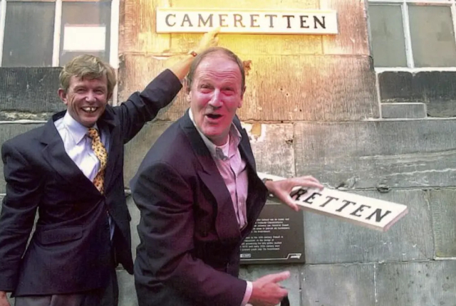 Twee lachende mannen houden een bord met 'RETTEN' vast.